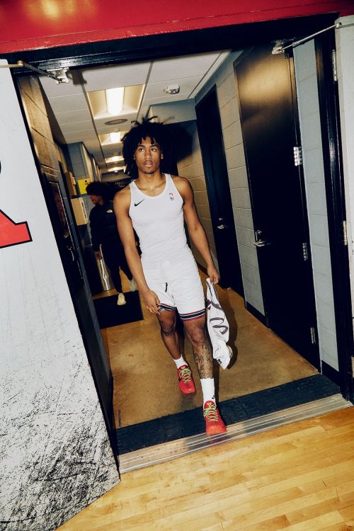 Young African-American male basketball player in a white uniform walks through a hallway, holding a towel and wearing red shoes.