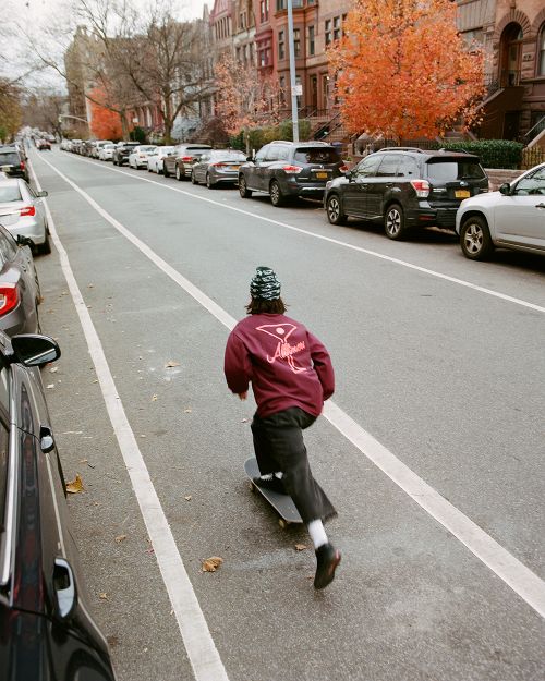 Person skateboarding on a street lined with parked cars and brick buildings during autumn, featuring trees with orange leaves.