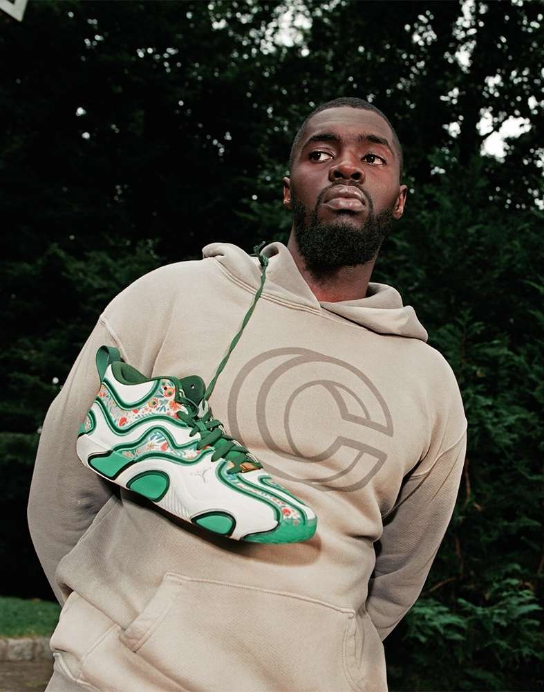 Stylish adult male wearing a tan hoodie with a logo, showcasing a white and green sneaker; outdoor setting with leafy trees in background.