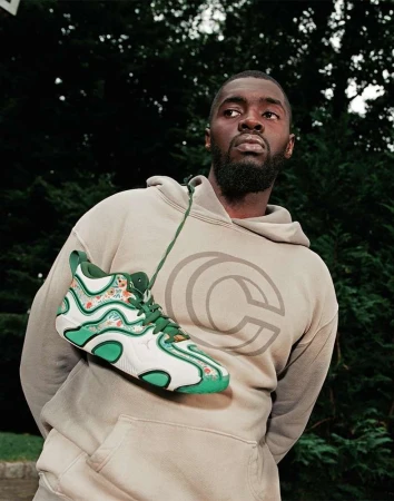 Stylish adult male wearing a tan hoodie with a logo, showcasing a white and green sneaker; outdoor setting with leafy trees in background.