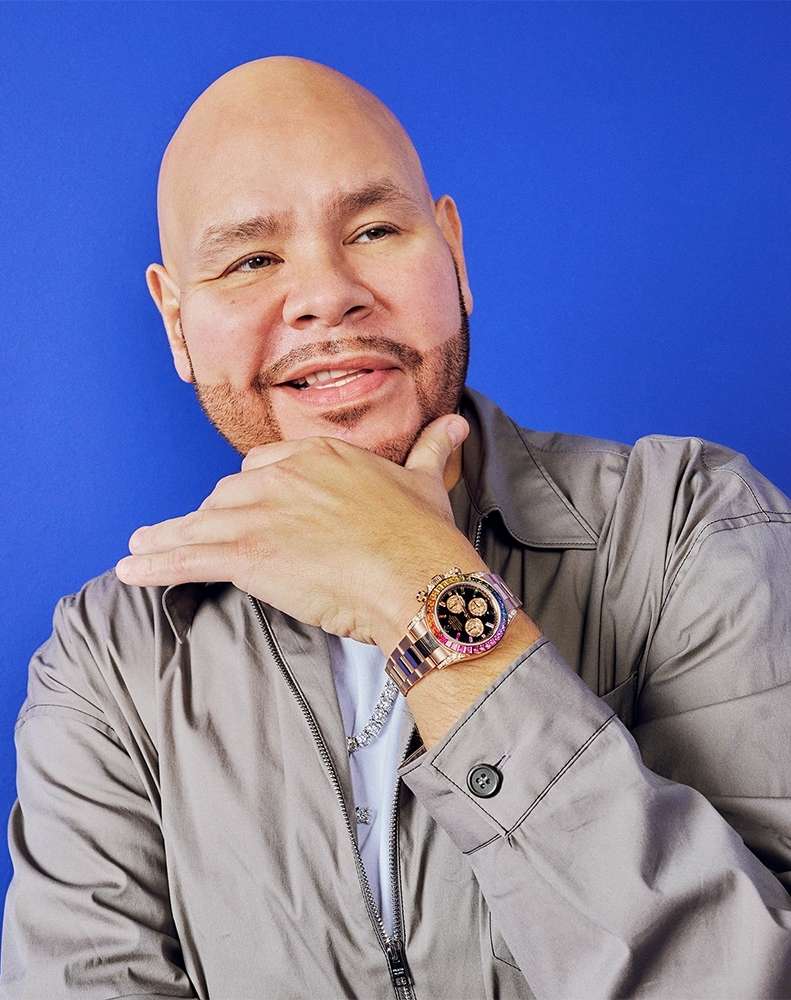 Adult male with a shaved head poses in front of a blue background, wearing a grey jacket, white t-shirt, and watch.
