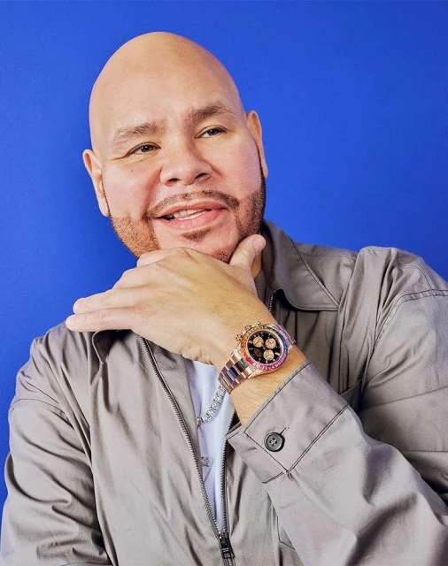 Adult male with a shaved head poses in front of a blue background, wearing a grey jacket, white t-shirt, and watch.
