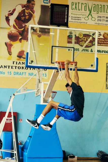 A man in athletic wear hangs from a basketball hoop in an indoor court, performing a pull-up, with gym equipment visible in background.