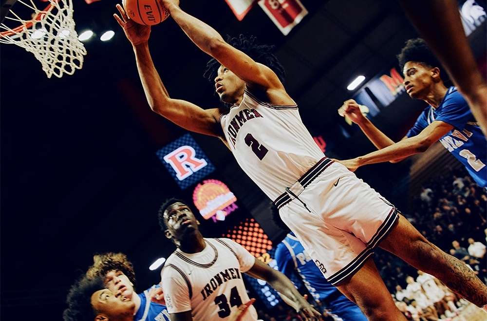 Basketball player in action, soaring toward the net during a game with other players and spectators in the arena.