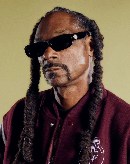 Portrait of Snoop Dogg wearing sunglasses and a varsity jacket, showcasing his braided hair against a yellow backdrop in a close-up studio shot.