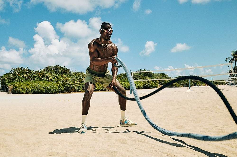 An adult Black man is doing a battle ropes exercise on a sandy beach with a volleyball net and greenery in the background on a sunny day.