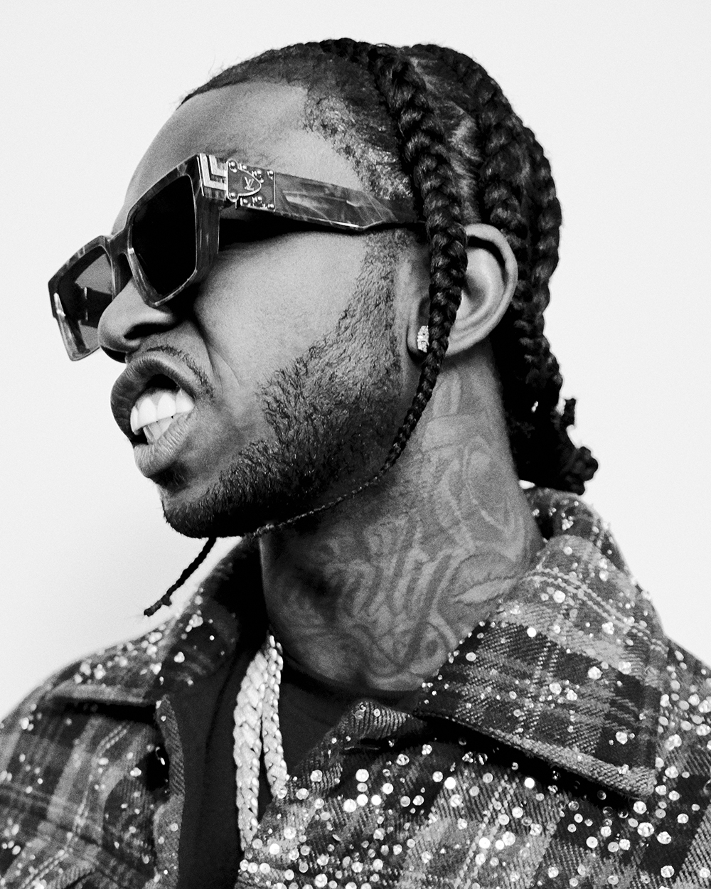 Black and white portrait of a man with cornrow braids wearing sunglasses, a patterned jacket, and jewelry against a plain background.