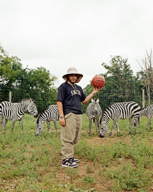 Adult male balances basketball in outdoor setting with zebras grazing in the background; he is wearing a large hat, baggy pants, and shoes.