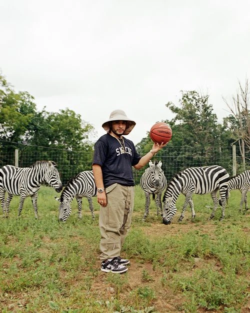 Adult male balances basketball in outdoor setting with zebras grazing in the background; he is wearing a large hat, baggy pants, and shoes.