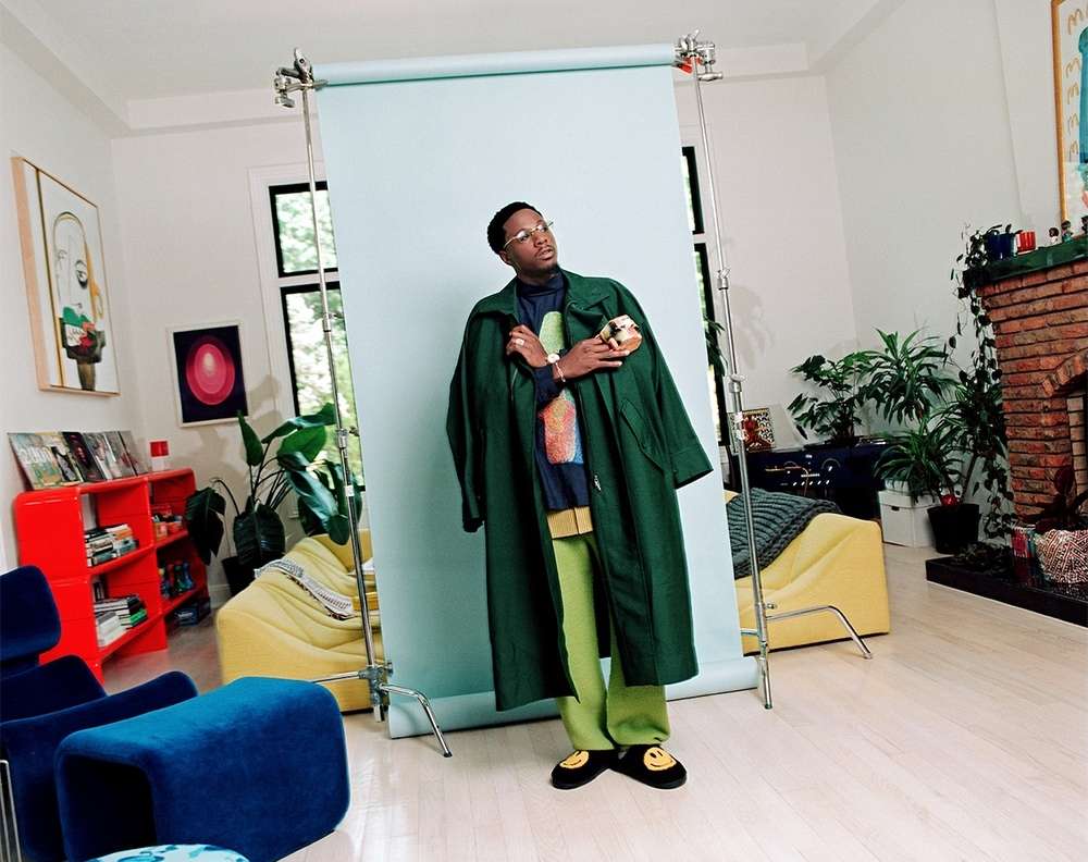 An African-American man in a green coat and lime pants stands in front of a pale-blue backdrop in a bright, naturally lit room.