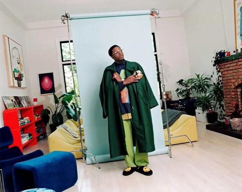 An African-American man in a green coat and lime pants stands in front of a pale-blue backdrop in a bright, naturally lit room.