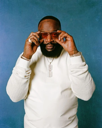 Rick Ross adjusting amber-tinted sunglasses, wearing a white sweatshirt and dog tag pendant against a blue textured backdrop.