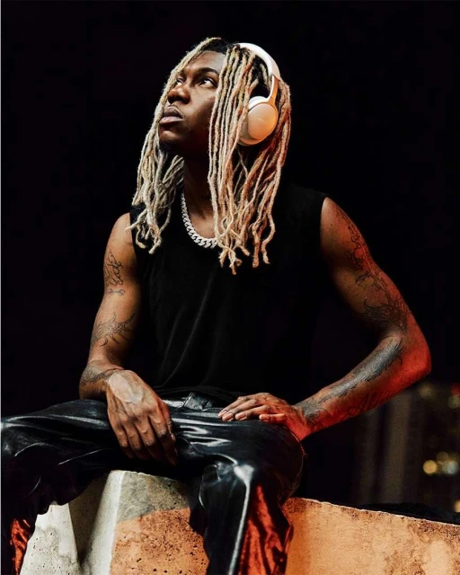 Portrait of a man with blonde dreadlocks wearing headphones and a sleeveless shirt sitting on a concrete wall at night.