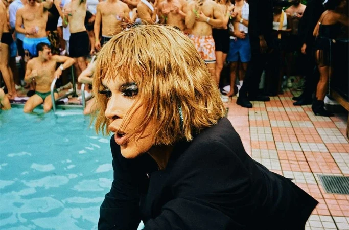 Person with blonde hair and black jacket near a pool surrounded by a group of people, creating a vibrant and lively atmosphere.