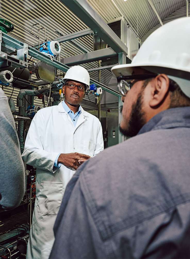 Two industrial workers in hard hats in a factory setting, one wearing a lab coat and glasses, discussing equipment.