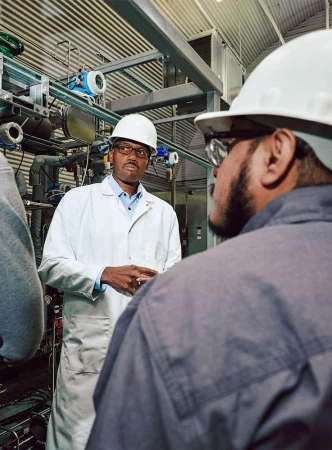Two industrial workers in hard hats in a factory setting, one wearing a lab coat and glasses, discussing equipment.