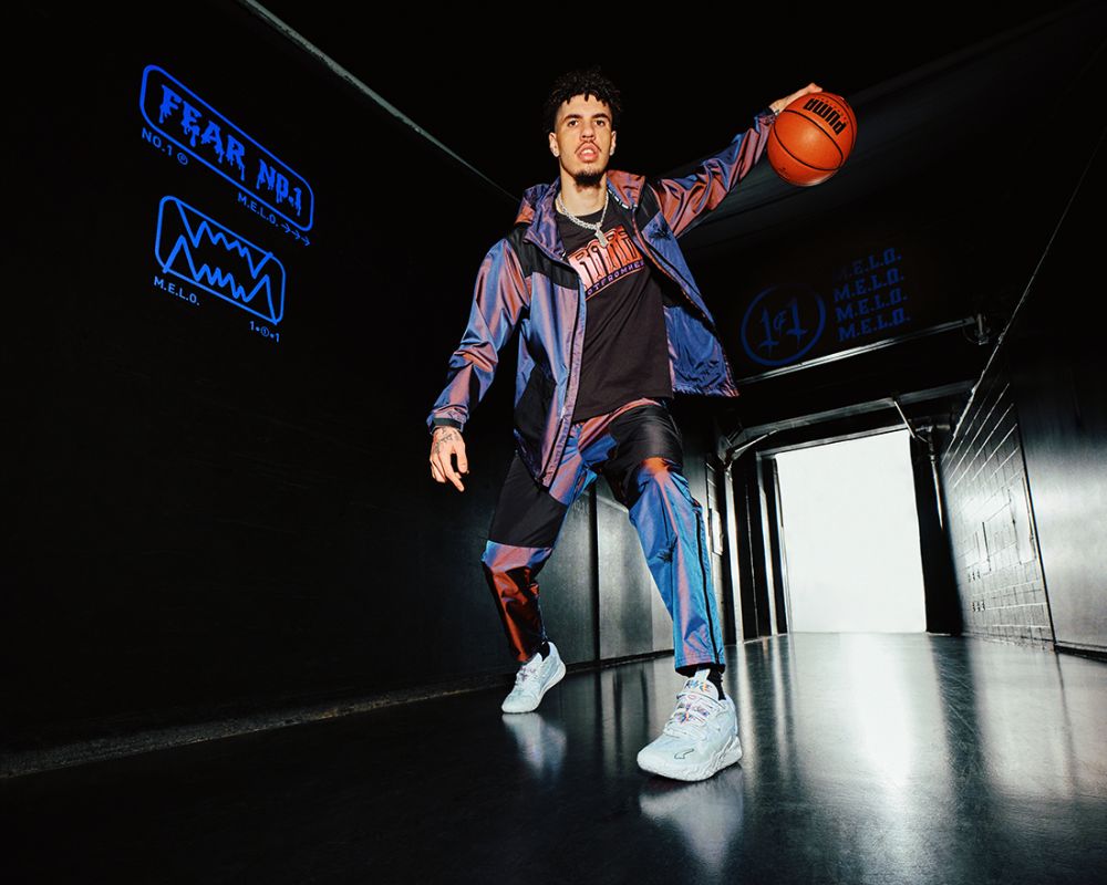 Lamelo Ball poses indoors with a basketball in sportswear, featuring a jacket, pants, t-shirt, and sneakers, against a dark background.