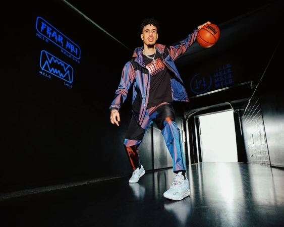 Lamelo Ball poses indoors with a basketball in sportswear, featuring a jacket, pants, t-shirt, and sneakers, against a dark background.