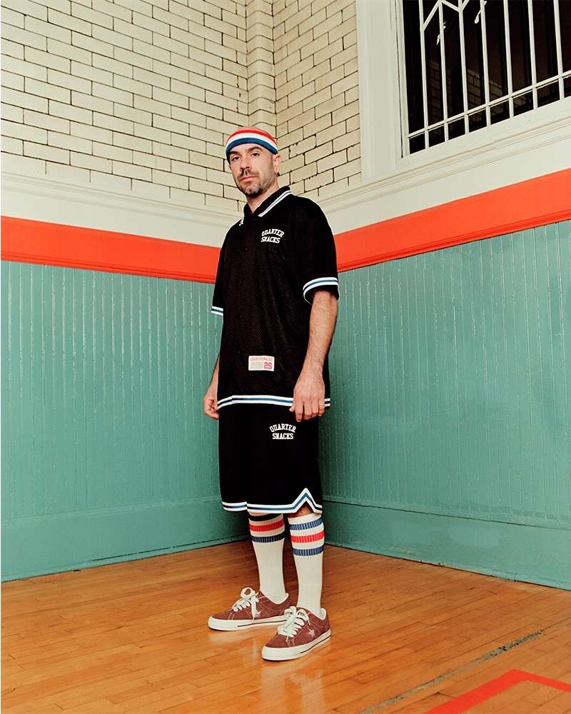 A man in a black jersey and shorts, styled with tube socks and sneakers, stands confidently on a basketball court with a vintage vibe.