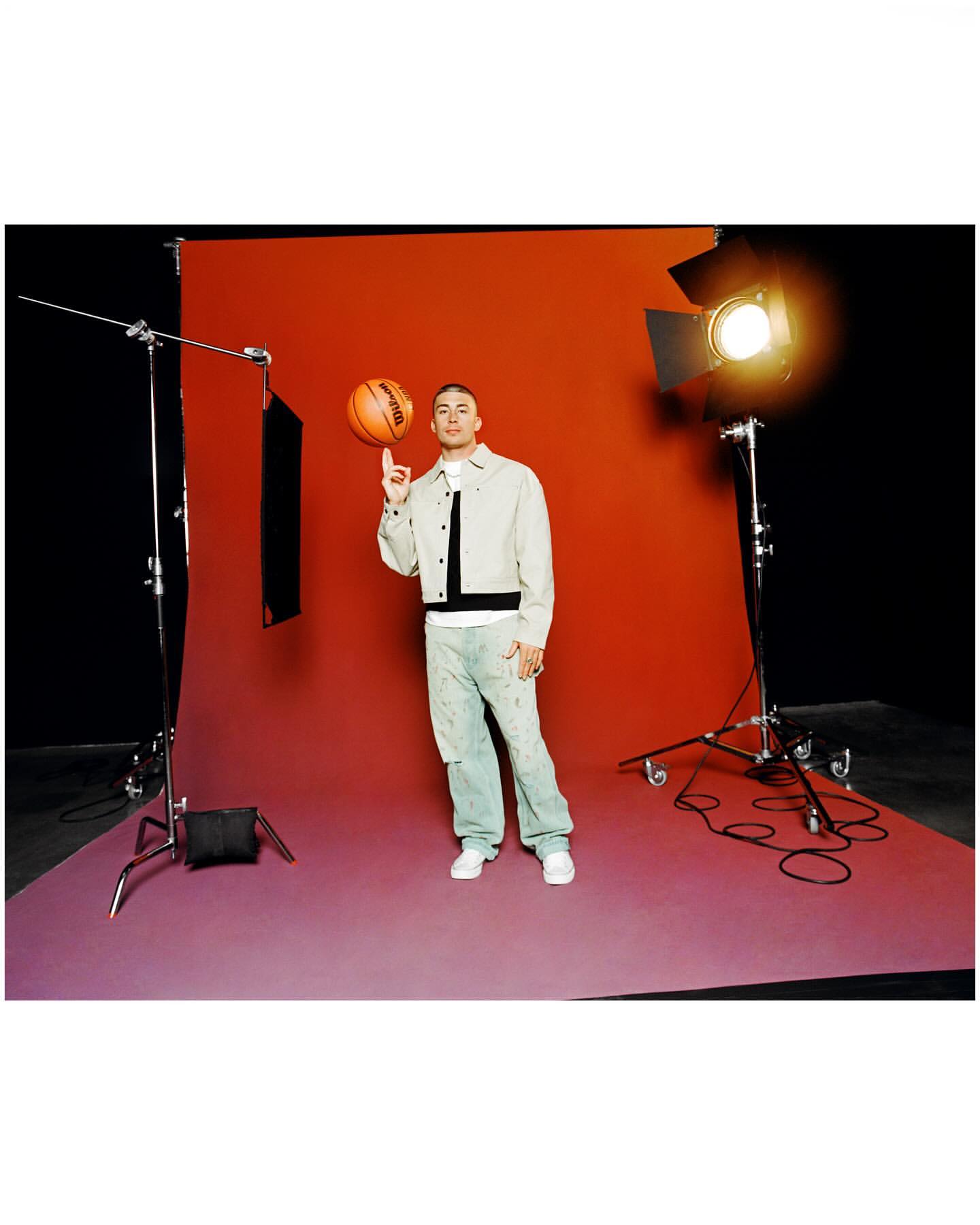 A man in a beige jacket spins a basketball on his finger in a studio setting with lighting equipment and an orange and black backdrop.