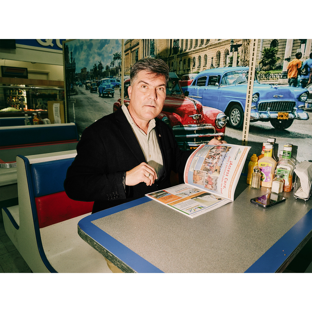 Man in a diner booth is reading a menu with vintage car mural in the background. He is wearing a suit jacket over a collared shirt.