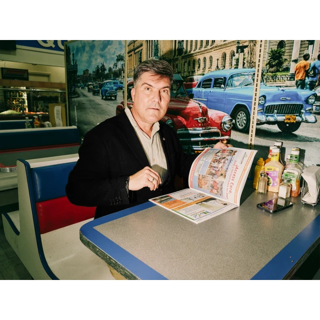 Man in a diner booth is reading a menu with vintage car mural in the background. He is wearing a suit jacket over a collared shirt.