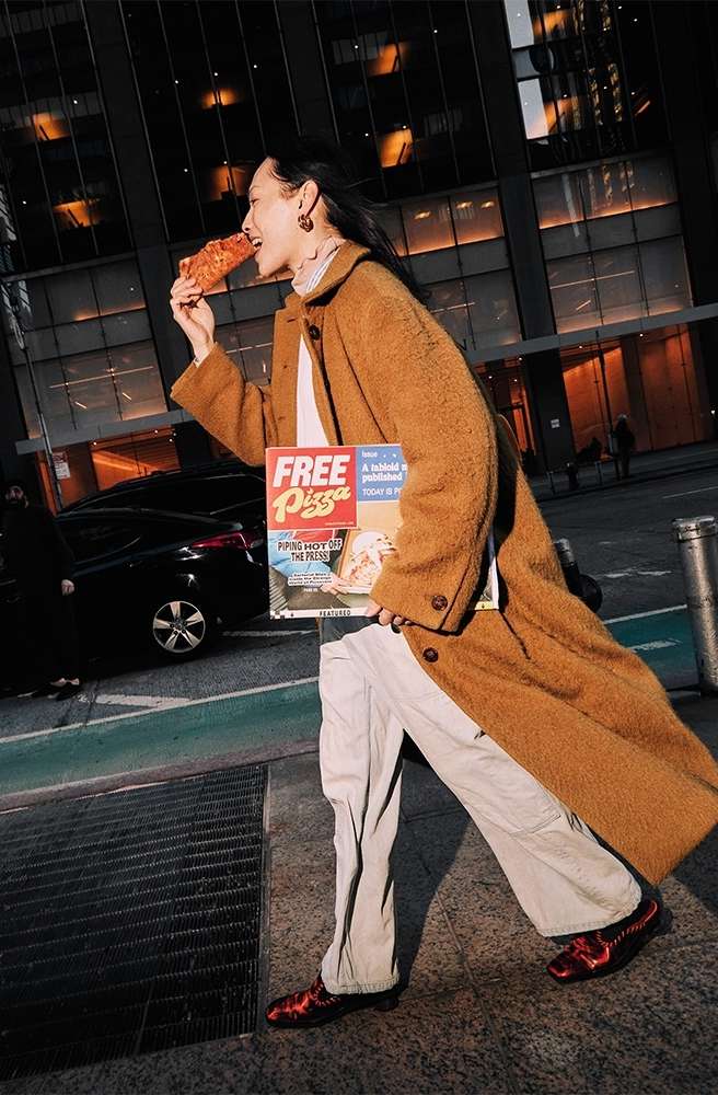 Woman in brown coat walking down a city street while eating pizza and holding a pizza box