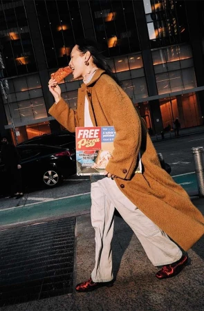 Woman in brown coat walking down a city street while eating pizza and holding a pizza box