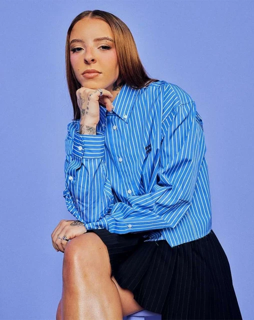 Portrait of a tattooed young woman with long auburn hair, wearing a blue-and-white striped shirt and black skirt, posing against a light purple backdrop.