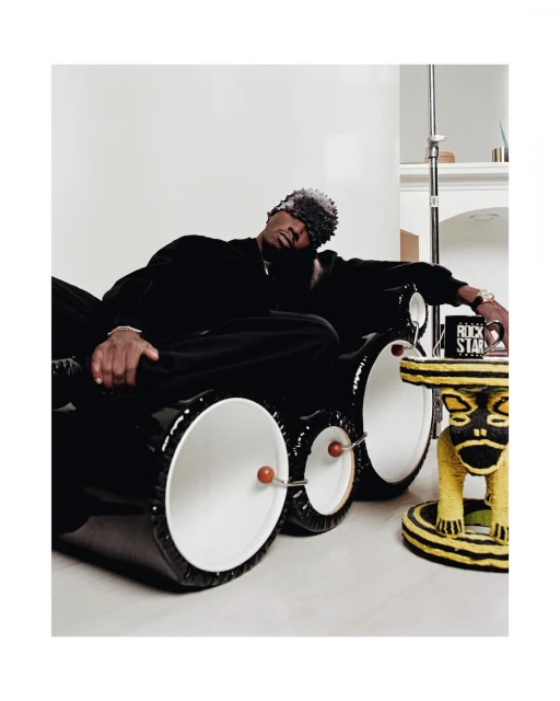 An adult male wearing black clothing rests on a black and white drum-shaped couch beside a yellow and black bear-shaped stool.