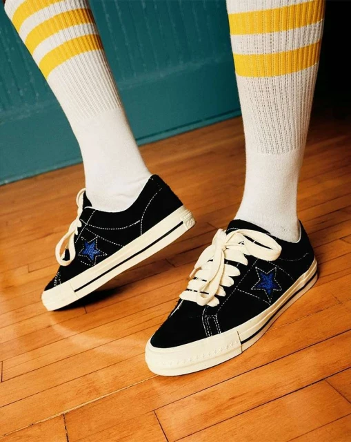 Close-up studio shot of black One Star Converse sneakers with white laces, cream soles, and white striped yellow socks on a wood floor.