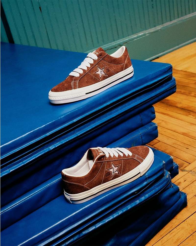 Two brown suede Converse One Star sneakers with white laces and soles stacked on a blue gym mat in a brightly lit indoor setting.