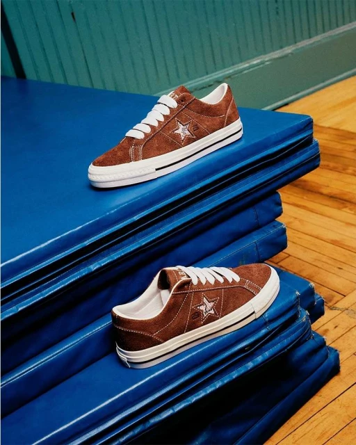 Two brown suede Converse One Star sneakers with white laces and soles stacked on a blue gym mat in a brightly lit indoor setting.