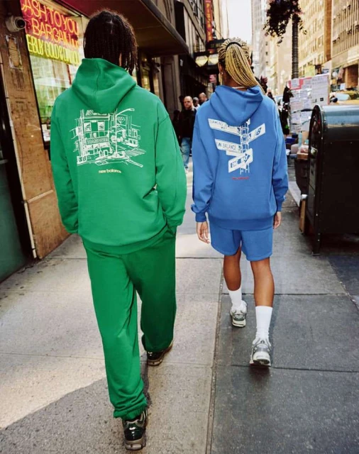 Two people wearing matching New Balance tracksuits walk on a city sidewalk. The person on the left wears a green tracksuit; the other wears blue.