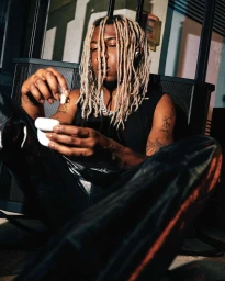 Black man with blonde dreadlocks seated outdoors holding wireless earbuds, photographed by David Cabrera