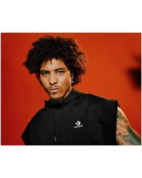 Portrait of a man with dark curly hair wearing a black sleeveless hoodie against a warm orange background.