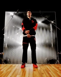 Low angle shot of a man wearing a black hoodie with red sleeves and black cargo pants standing before a translucent backdrop.