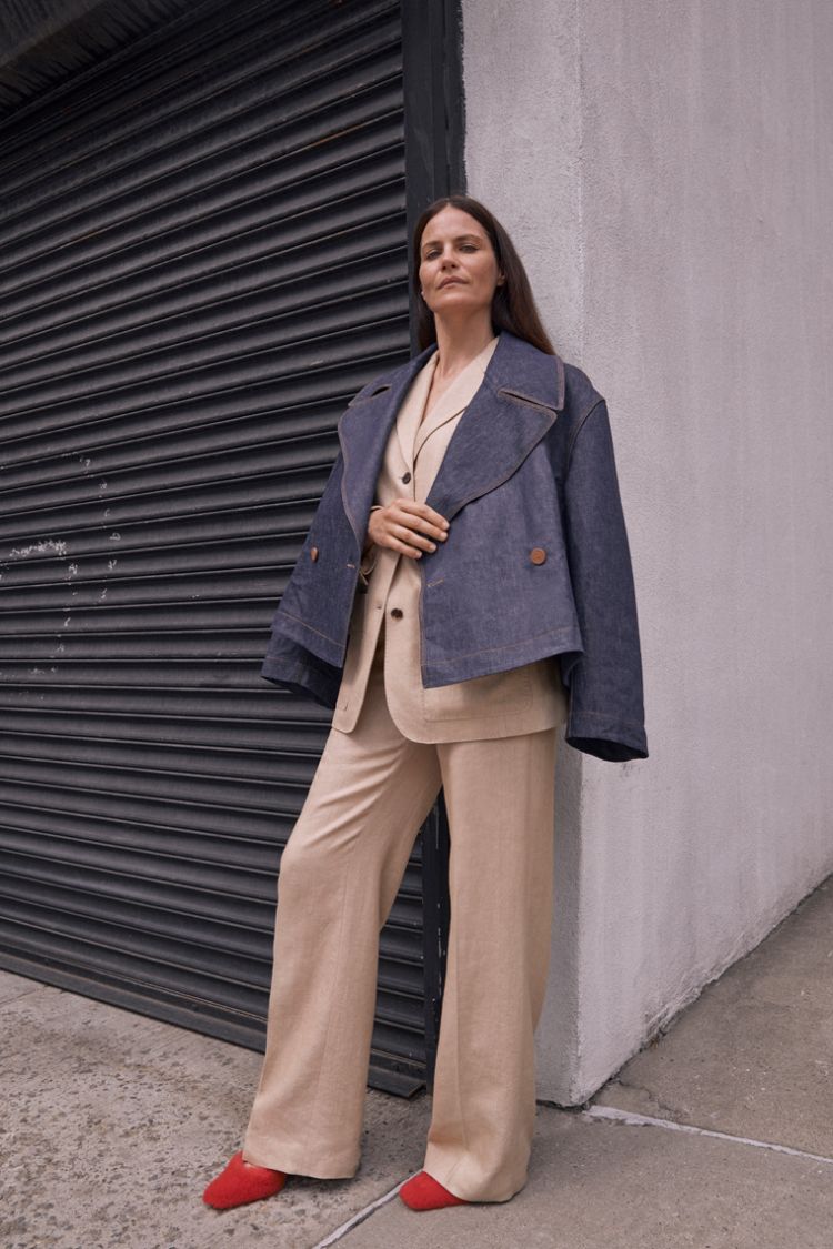 Adult female wearing a denim jacket and beige suit standing in front of a gray corrugated metal door and a light gray wall.