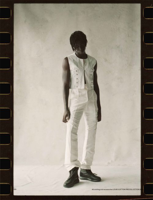 An African American model poses in a photo studio wearing a white vest, white pants, and black boots, framed by filmstrip edging.