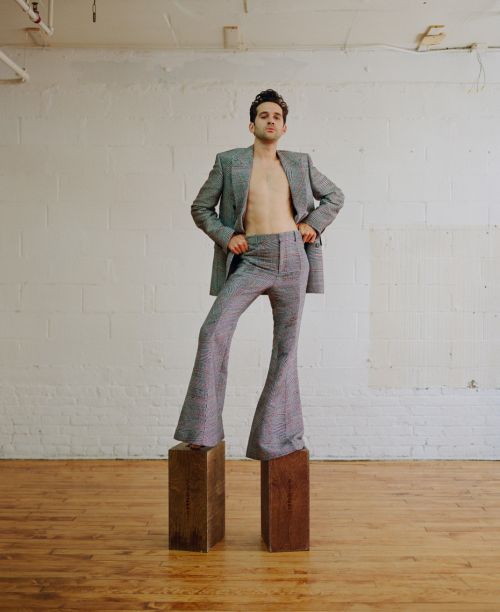 A young adult male in a grey tweed suit strikes a confident pose atop wooden blocks in a photo studio, set against a stark white wall.