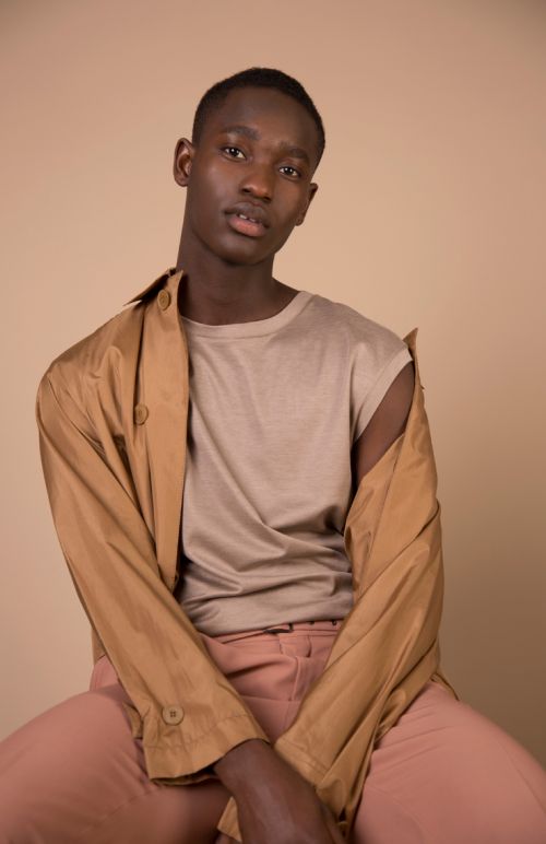 Portrait of a dark-skinned African male in a light-beige t-shirt and peach-colored pants with a brown jacket draped over his shoulders.