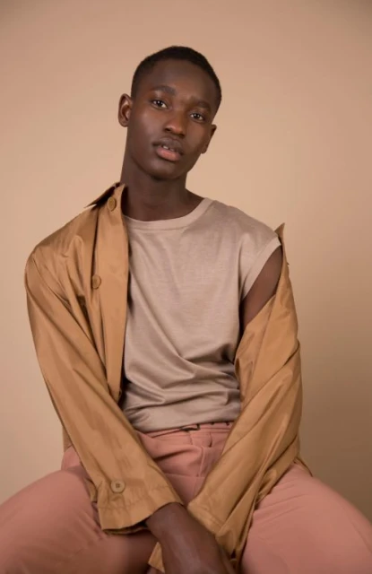 Portrait of a dark-skinned African male in a light-beige t-shirt and peach-colored pants with a brown jacket draped over his shoulders.