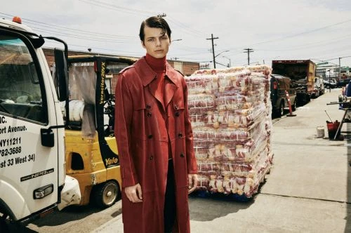 Fashion shot of a male model in a red trench coat standing on a city street with trucks and large food pallets, against a clear sky.