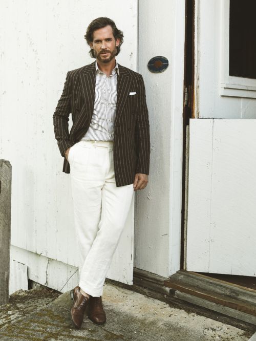 Man in striped blazer, white pants, and brown boots poses confidently against a white wall, exuding classic style and sophistication.