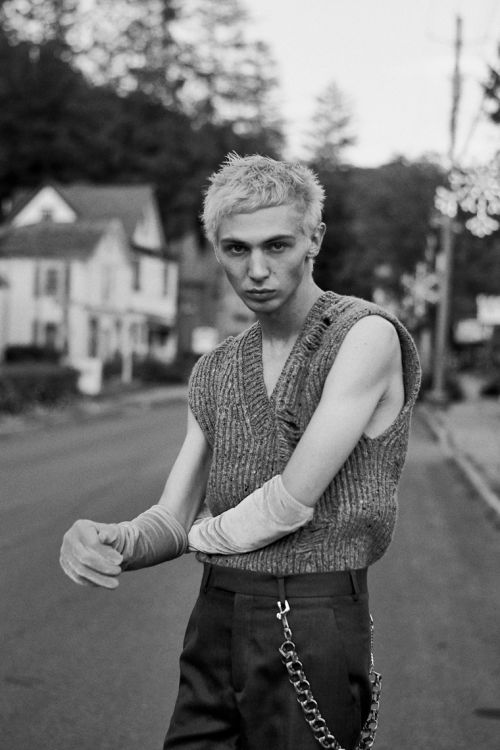 Black and white portrait of a young adult male with bleached hair, wearing a knit vest and gloves, posing on a residential street.