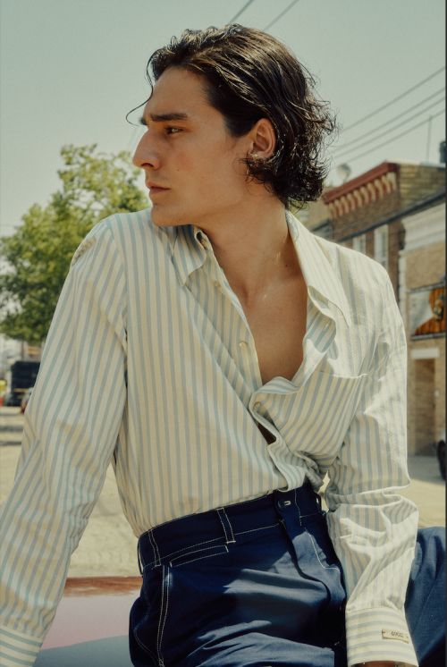 A young adult man with dark hair wearing a striped shirt and dark pants is photographed outdoors in daylight against a building backdrop.