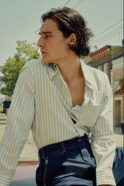 A young adult man with dark hair wearing a striped shirt and dark pants is photographed outdoors in daylight against a building backdrop.
