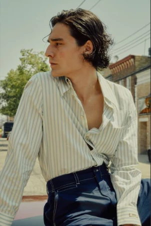 A young adult man with dark hair wearing a striped shirt and dark pants is photographed outdoors in daylight against a building backdrop.