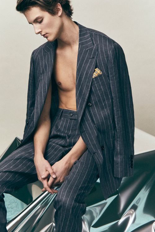 An adult male model poses in a pinstripe suit on a silver reflective surface, showcasing fashion, style, and contemporary menswear.
