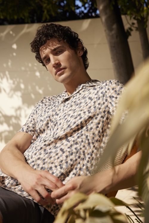 Handsome man in a patterned shirt poses outdoors in dappled sunlight, showcasing a blend of casual style and natural lighting.